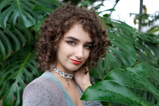 Woman with curly hair is wearing a necklace and a red lipstick. She is standing in front of a green plant