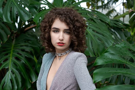 Woman with curly hair is wearing a necklace and a red lipstick. She is standing in front of a green plant