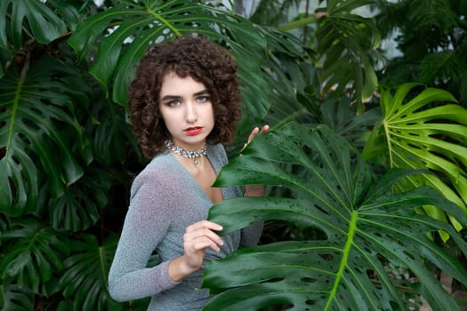 Woman is standing in front of a large leafy plant. She is wearing a silver necklace and a red lipstick