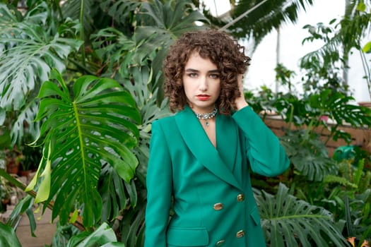 Woman wearing a green jacket and a necklace. She is standing in front of a plant. The jacket is green and has a collar