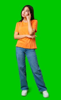 A young woman dressed in an orange shirt and blue jeans stands with one hand on her chin, appearing to contemplate or think deeply. The vivid green background adds focus to her expression.
