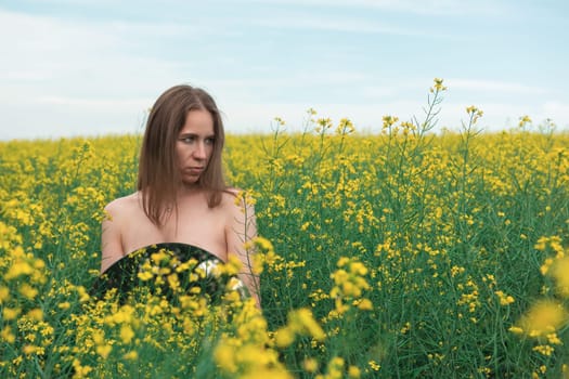A young woman stands in a vast yellow flower field, holding a round mirror. She gazes calmly, blending with the serene natural scenery.