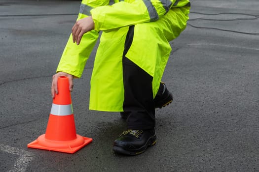 A worker wearing a bright yellow high-visibility raincoat and safety boots places an orange traffic cone on a wet asphalt road.