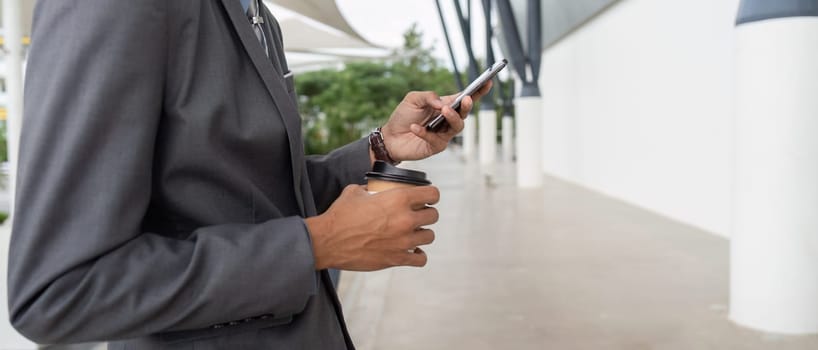 A businessman in a suit holding a coffee cup and checking his smartphone while standing outdoors in a modern setting, combining work and refreshment.