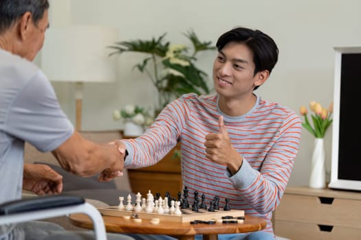 A young man smiling and giving a thumbs up while shaking hands with his father after an enjoyable chess game at home, celebrating family moments.