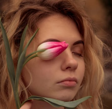 very close-up of a blonde with a flower near her face. tulip she has one eye. portrait of a girl, near her face she holds one pink tulip. dressed in a beige coat. she has light long wavy hair.