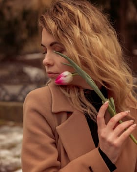 close-up of a blonde with a flower near her face.portrait of a girl, near her face she holds one pink tulip. dressed in a beige coat. she has light long wavy hair. she is thoughtful, it is warm spring