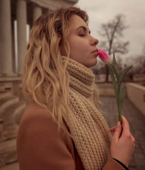 close-up of a blonde with a flower near her lips. portrait of a girl, near her face she holds one pink tulip. she stands against the background of an 18th century colonnade. dressed in a beige coat