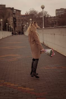 Blonde with a bouquet of pink tulips in the city in full growth. The girl dances with developing curls in the wind. She looks into the frame and smiles. She is dressed in a beige classic coat