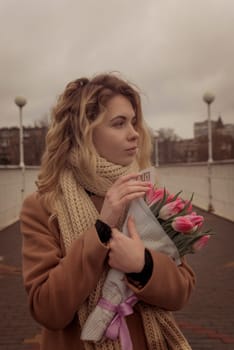 Close-up of a blonde with a bouquet of pink tulips. She is walking around the city, smiling, her hair is flying. She is wearing a beige classic coat. And a warm scarf. The girl is looking away