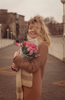 Blonde with a bouquet of pink tulips in the city. Girl with curls developing in the wind. She looks into the frame and smiles. Dressed in a beige classic coat. And a warm scarf, large knit.