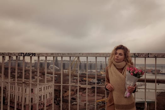 Blonde with a bouquet of pink tulips standing on the bridge. Behind her is a seaport. She is looking away. She is wearing a beige classic coat. And a warm, large-knit scarf.