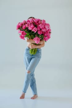 A woman in jeans holds a huge bouquet of pink peonies, completely hiding her upper body. The minimalist background, barefoot pose, and bright flowers create a stylish, artistic, and modern aesthetic.