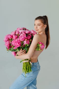 A woman in jeans holds a huge bouquet of pink peonies, completely hiding her upper body. The minimalist background, barefoot pose, and bright flowers create a stylish, artistic, and modern aesthetic.