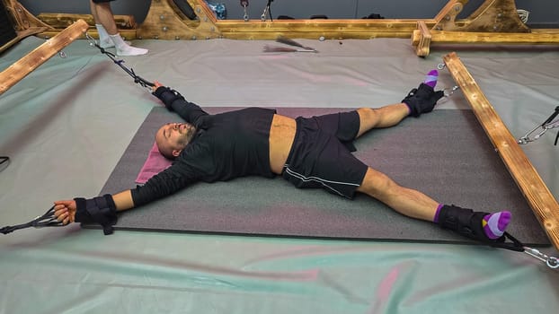Man undergoing physiotherapy treatment, lying on a mat with arms and legs outstretched, attached to suspension straps