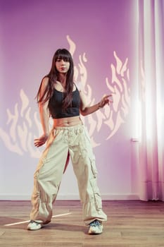 A woman performs an expressive dance routine in a spacious studio. She wears loose beige pants and a black top, moving confidently under soft, colorful lights that enhance the atmosphere.