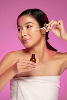 A young woman with long dark hair is confidently applying a serum to her face. She is clothed in a white towel and exudes a sense of enjoyment in her skincare ritual against a vibrant pink backdrop.