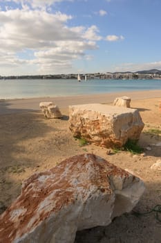A serene beach with large rocks, calm waters and a town visible on the other side of the bay, portocolom.mallorca, balearic islands, spain
