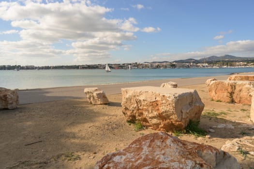 Beach scene with large rocks and a sailboat visible across the calm water, portocolom.mallorca, balearic islands, spain