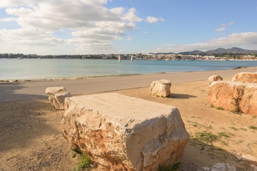 Beach with a distant view of the town and sailboat under a partly cloudy sky, portocolom.mallorca, balearic islands, spain.