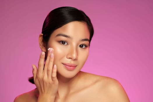 A young woman gently applies a skincare product to her cheek while looking at the camera. She has a serene expression and is set against a vibrant pink backdrop, showcasing her beauty routine.