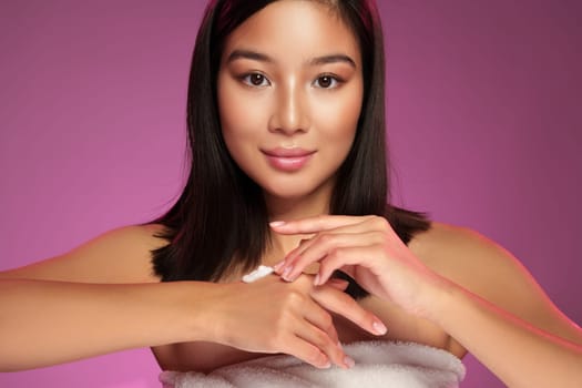 A young woman is applying skin cream to her arms while looking at the camera with a gentle smile. The vibrant pink background highlights her clear skin and beauty routine.