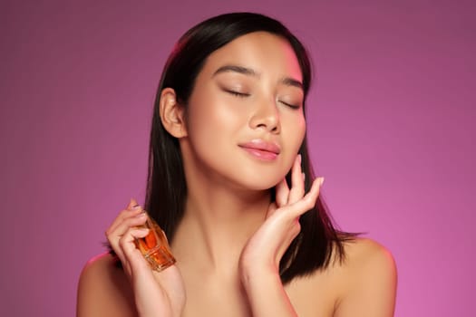 A young woman with straight black hair gently holds a small bottle of perfume near her face, smiling softly with her eyes closed. Her skin glows under the pink light, reflecting tranquility.