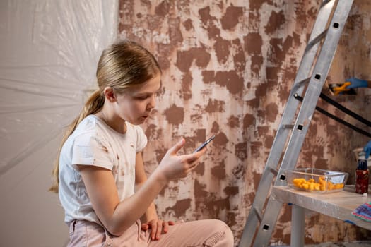 A focused young girl sits in a creative setting, engaging with her phone and surrounded by art supplies.