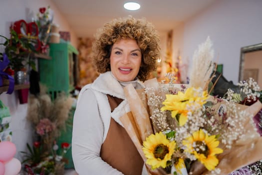 A woman with curly hair smiles brightly as she holds a beautiful bouquet filled with sunflowers and delicate foliage. She stands in a cozy floral shop adorned with colorful arrangements.