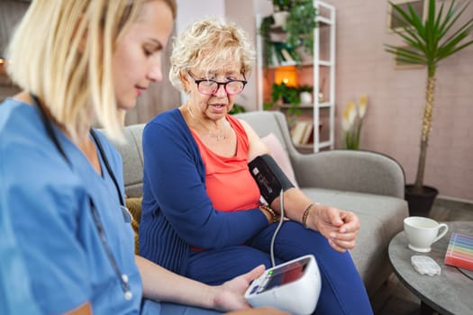 Caring nurse measures blood pressure of senior woman in cozy living room