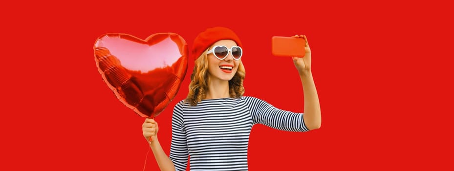 Summer portrait of happy smiling young woman taking selfie with smartphone holding red heart shaped balloon wearing french beret, sunglasses isolated on studio background