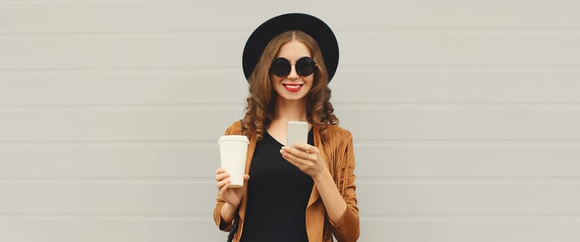 Stylish happy smiling young woman with phone, beautiful girl holding smartphone, cup of coffee wearing black hat standing on city street on gray background