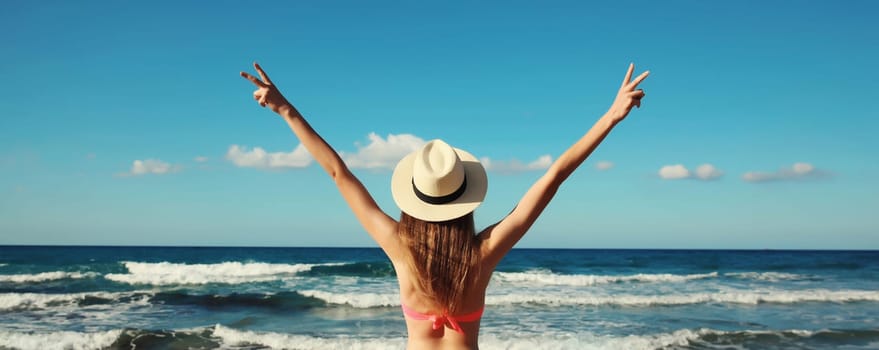 Summer vacation, rear view of beautiful happy slim woman in bikini swimsuit and tourist straw hat raising her hands up on the beach on sunny day on sea coast with waves