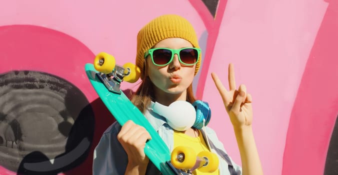 Stylish young girl with skateboard in hat, glasses, listening to music with headphones, modern teenager girl posing against the background of pink graffiti wall