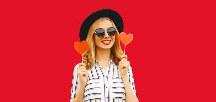 Portrait of happy cheerful young woman with sweet red heart shaped lollipop on stick wearing a black round hat isolated on studio background
