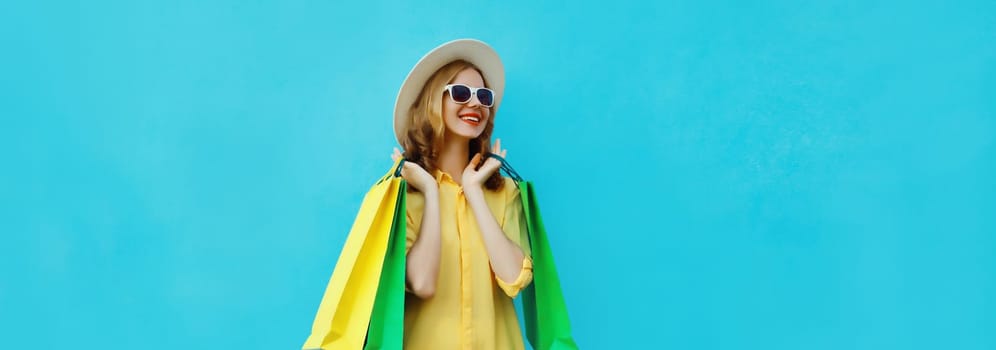 Fashion, purchase. Stylish beautiful happy young woman posing with colorful shopping bags, girl in summer hat looking away, standing on bright blue background