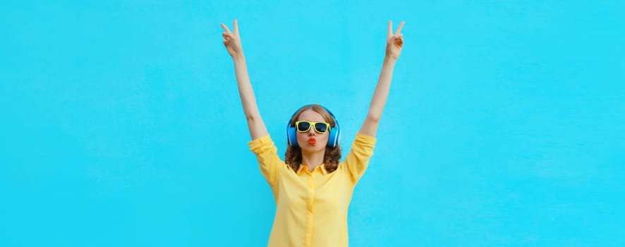 Happy modern young woman dancing listening to music with wireless headphones having fun raises her hands up isolated on colorful blue studio background