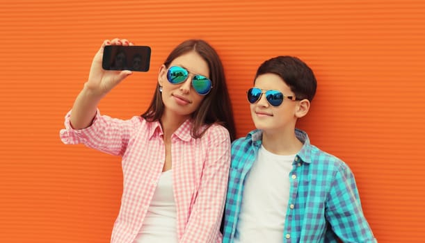 Happy mother with son teenager taking selfie with smartphone wearing summer glasses in the city on orange wall background