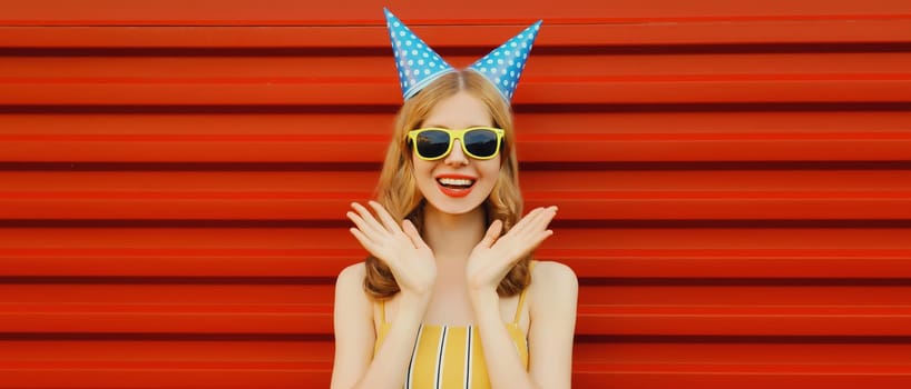 Holiday, celebration, event. Happy cheerful young woman laughs celebrating having fun at a party wearing festive birthday hat on red background