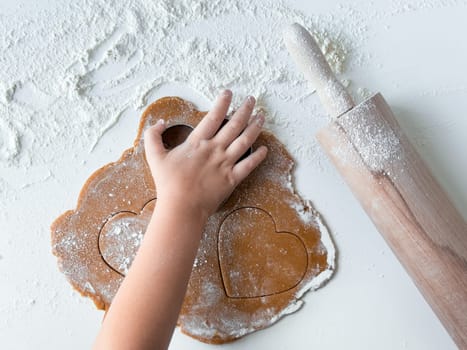 Child making heart shaped cookies with rolling pin on floured surface.