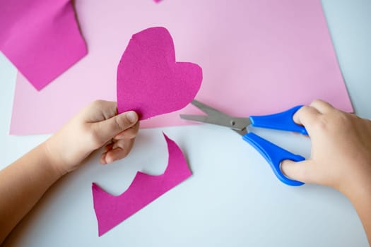 Child creating pink paper heart with blue scissors on craft project.