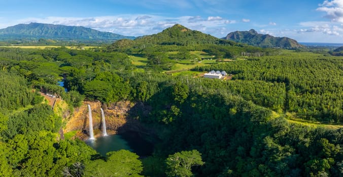 Aerial view of Wailua Falls cascading into a green valley on Kauai Island, Hawaii, surrounded by tropical vegetation, hills, and distant mountains.