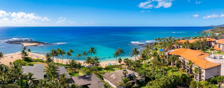 Aerial view of Kauai island's tropical coastline featuring a sandy beach, turquoise waters, palm trees, and residential buildings with orange tiled roofs.