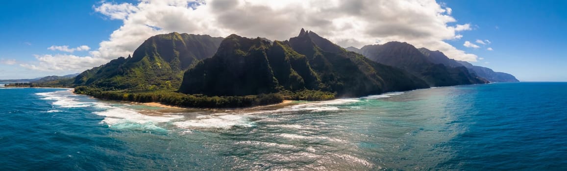 Aerial view of jagged, green covered Napali Coast cliffs on Kauai Island, Hawaii, with turquoise ocean, sandy beaches, and sunlight through clouds.