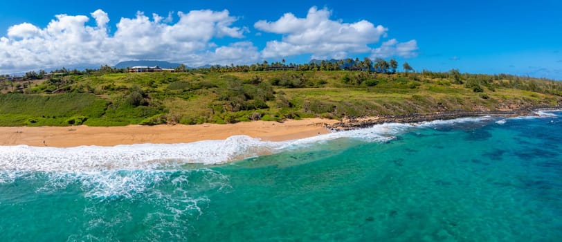 Aerial view of a golden sandy beach on Kauai Island, Hawaii, bordered by turquoise waters and lush greenery, with scattered trees and distant buildings.