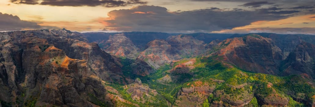 Aerial view of Waimea Canyon on Kauai Island, Hawaii, featuring rugged cliffs in red, orange, and green hues, with lush vegetation and soft clouds above.