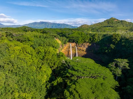 Twin streams of Wailua Falls cascade into a hidden pool, surrounded by dense tropical vegetation and rugged mountains under a clear blue sky.