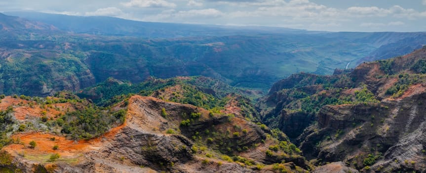 Aerial perspective of Waimea Canyon on Kauai Island, Hawaii, featuring red and green cliffs, rugged terrain, deep valleys, and a partly cloudy sky.