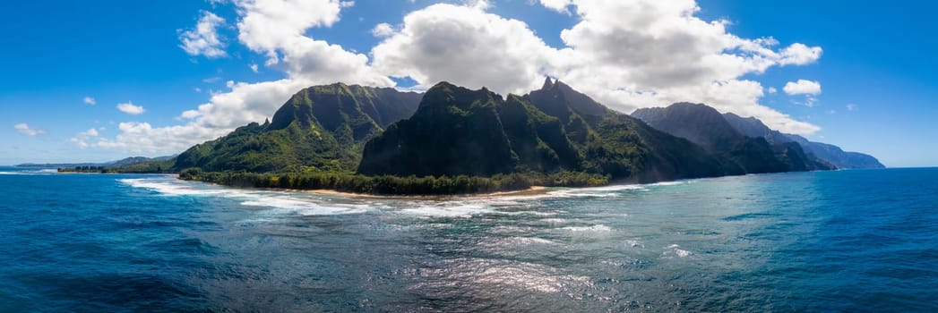 Aerial view of Kauai's Napali Coast with jagged green cliffs, sandy beach, turquoise ocean waves, and a partly cloudy sky creating vivid contrasts.