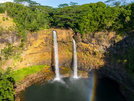 Twin waterfalls of Wailua Falls on Kauai, Hawaii, cascade from a rocky cliff into a pool, surrounded by lush greenery with a rainbow at the base.
